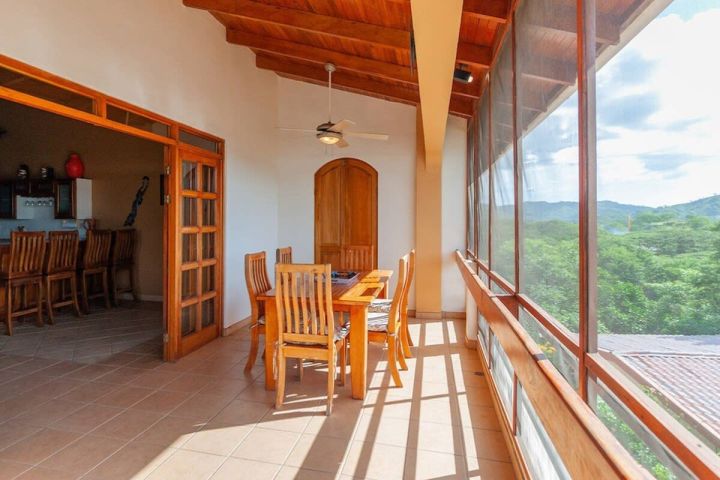 Sunny balcony with wooden dining set, ceiling fan, and a view of trees.