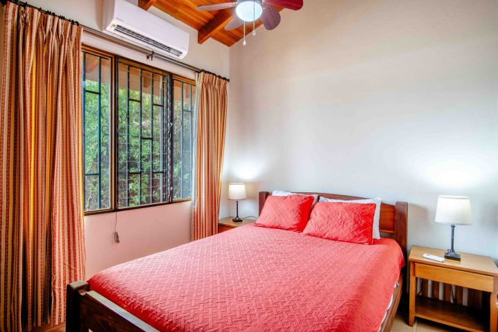 Bedroom with a red bedspread, two lamps, wooden ceiling, and a window with curtains.
