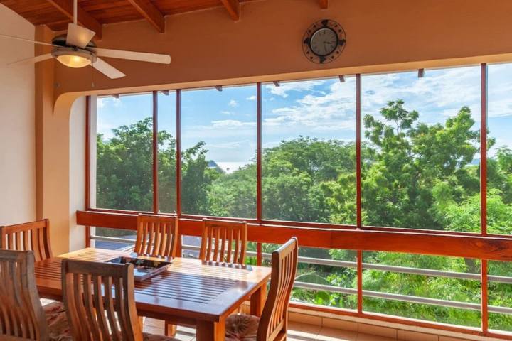 Dining area with wooden table, chairs, ceiling fan, large windows overlooking trees.