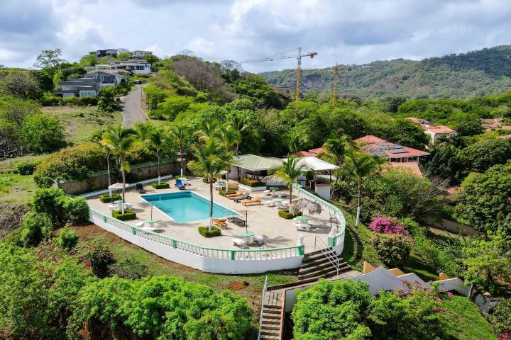 Aerial view of a villa with a pool and palm trees, surrounded by greenery and distant hills.
