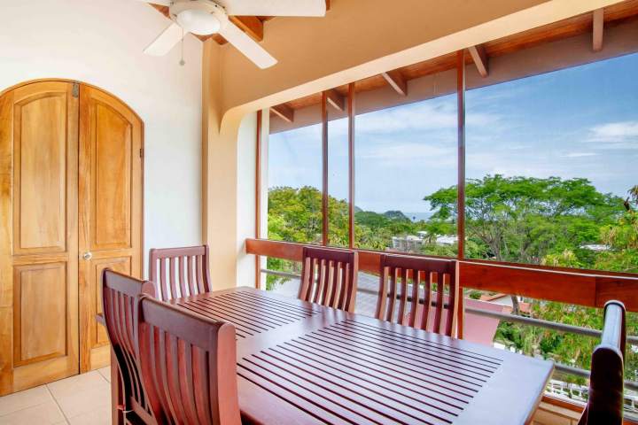 Dining table on balcony with wooden chairs, ceiling fan, and ocean view.