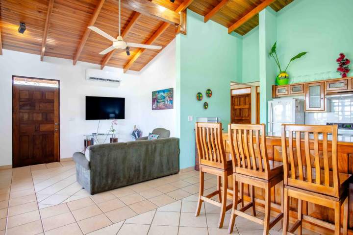 Living room with sofa, wall-mounted TV, wooden ceiling, and dining table with chairs.