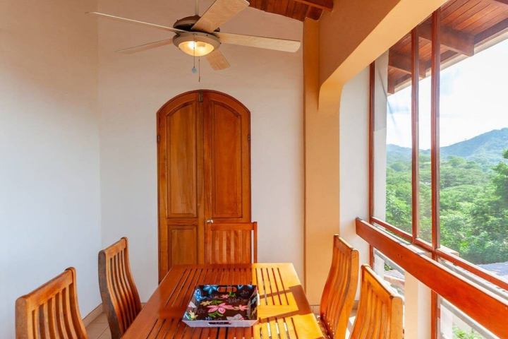 Dining area with wooden table, chairs, ceiling fan, wooden door, and large windows revealing a scenic view.