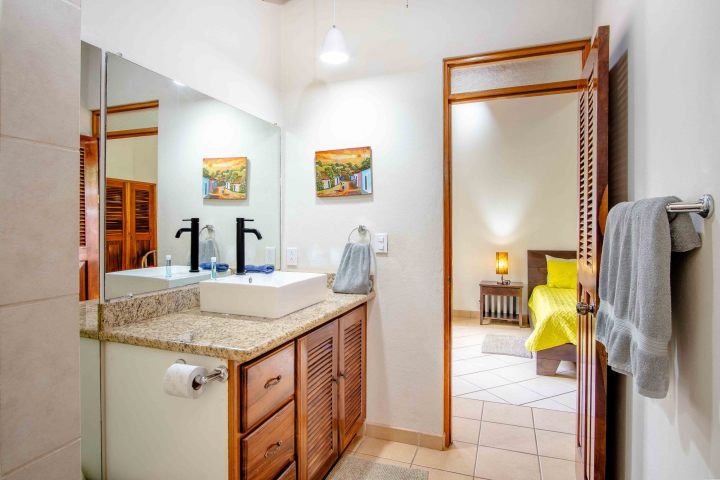 Bathroom with dual sinks, wooden cabinets, artwork on wall, view of bedroom with yellow bedding.