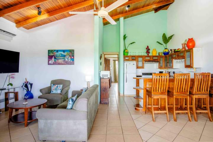 Cozy living room with sofa, kitchen, wooden chairs, and ceiling fan.